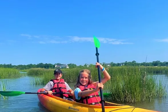 Two kids paddling a double kayak through marsh grass