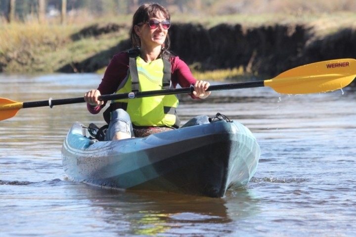 Person kayaking on a calm river, wearing a yellow life jacket and holding an orange paddle.