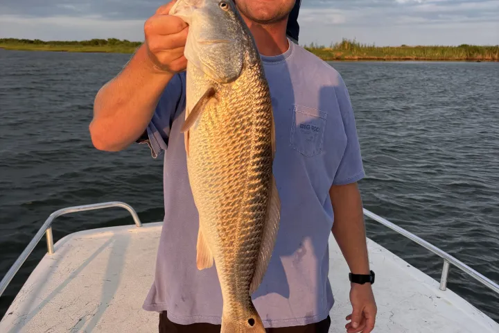 Two men on a boat, one holding a large fish, both wearing sunglasses, under a cloudy sky.