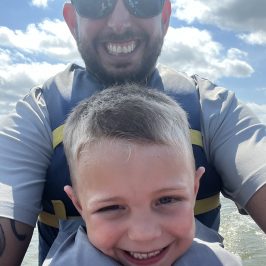 Man and child with life jackets smiling outdoors under a cloudy sky.