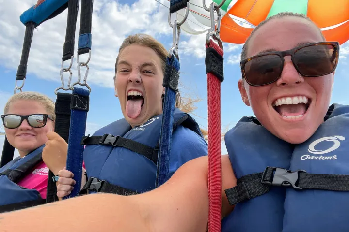 Three people parasailing with colorful parachute, smiling, wearing blue life vests and sunglasses.
